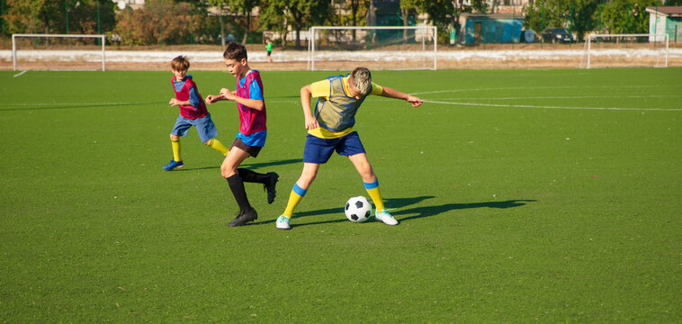 The photo features three young soccer players on a green artificial turf field,actively dribbling a soccer ball.They wear colorful jerseys and athletic gear, participating in a sports practice session