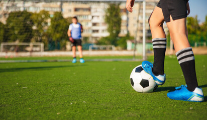 The photo shows a close-up of a soccer player preparing to kick the ball on a green turf field. In the background, another player is seen standing, ready for action.