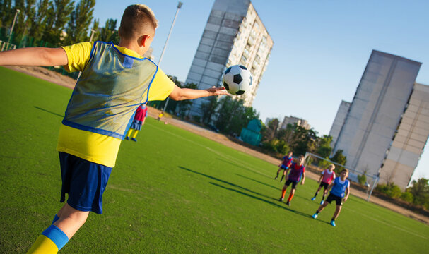 A young soccer player in a yellow shirt and blue shorts performs a throw-in while standing on a green field. Other players in colorful vests are in the background, with buildings visible behind.