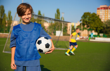 A young soccer player in a blue training vest holds a soccer ball, smiling at the camera. In the background, another player in a yellow shirt is in motion, with a goalpost and clear skies visible.
