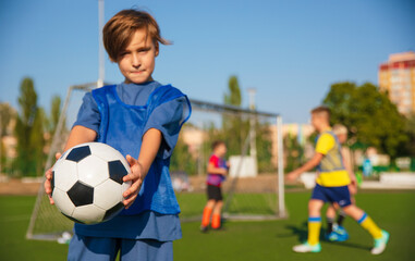 A young soccer player in a blue training vest holds a soccer ball, smiling at the camera. In the background, other players in yellow and purple vests are seen practicing on a green field.
