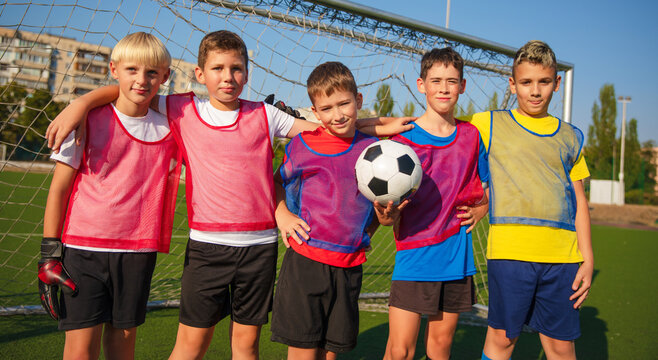 The photo shows five young soccer players posing in front of a goal, smiling and standing close together. They wear colorful jerseys, with one holding a soccer ball and another in goalkeeper gloves.