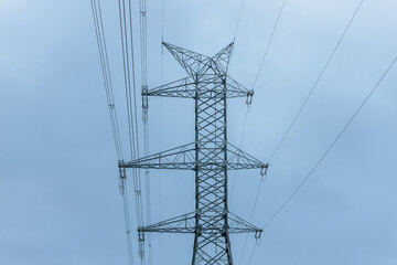 A towering high-voltage electricity pylon stands against a cloudy blue sky, supporting multiple power lines. The steel lattice structure highlights industrial energy infrastructure.