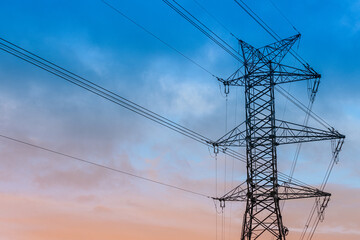 Fototapeta premium A high-voltage electricity pylon with power lines stretching across the sky during dusk. The silhouette contrasts with the gradient hues of blue and orange in the background.