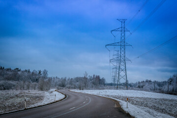 High-voltage electricity pylons stretch across a snowy landscape alongside a winding road under a blue winter sky. The scene captures the contrast between industrial infrastructure and nature.
