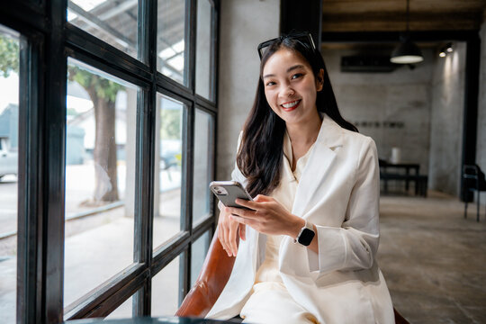 Young asian businesswoman is smiling and using a smartphone while sitting in a modern cafe - Powered by Adobe