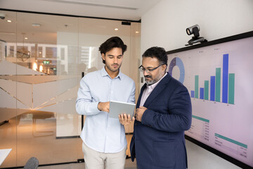 Young Latin employee showing electronic report to elder boss, consulting mature expert, asking...