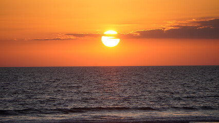The sun sets over the vast ocean, casting a warm glow across the sky and water. Gentle waves ripple towards the shore as clouds add depth to the breathtaking scene.

