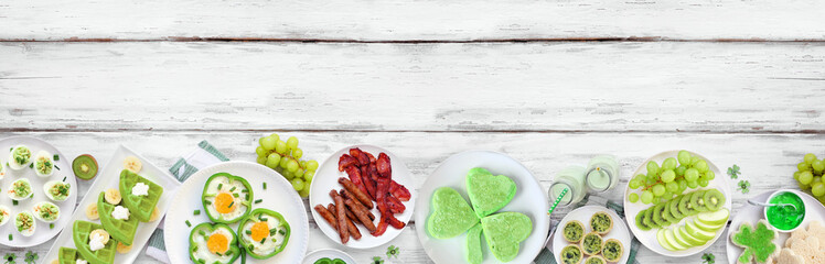St Patricks Day breakfast or brunch bottom border against a white wood banner background. Variety of green and shamrock themed pancakes, toasts, eggs, fruits and meats. Top view. Copy space.