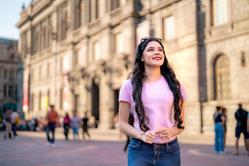 Happy young Latina woman with wavy hair, pink t-shirt, jeans, and a red backpack walks in front of the MUNAL museum in Mexico City, enjoying a sunny day and historic architecture.  