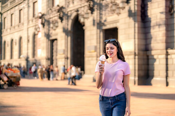 Obraz premium Happy young Latina woman with wavy hair, pink t-shirt, and jeans enjoys an ice cream cone while walking in front of the MUNAL museum in Mexico City on a sunny day. 