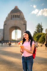Fototapeta premium Smiling young Latina woman with wavy hair, pink t-shirt, jeans, and a red backpack enjoys spicy mango in front of the Monument to the Revolution in Mexico City during a sunny afternoon. 