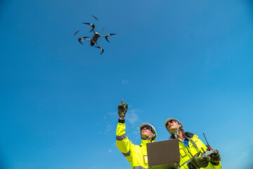 Two engineers in high visibility jackets and helmets observing a drone flying over a construction...