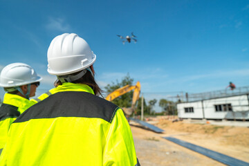 Obraz premium Two engineers in high visibility jackets and helmets observing a drone flying over a construction site, with machinery and modular buildings, highlighting technology and site monitoring.