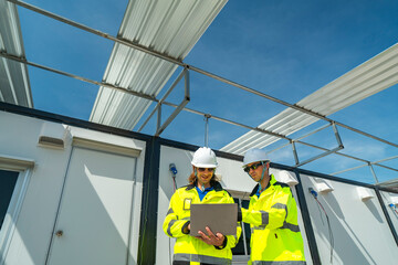Two engineers in high visibility jackets and helmets working on a laptop at a modular construction...