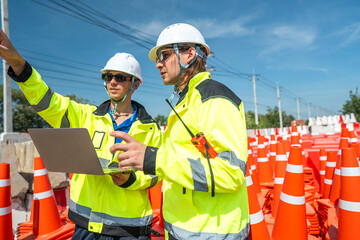 Two engineers in high visibility jackets discussing site plans on a laptop near a stack of orange traffic cones and concrete barriers, highlighting teamwork, safety, and project coordination.