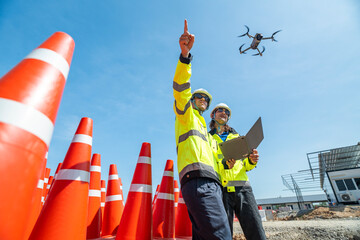 Two engineers in high visibility jackets and helmets operate a drone using a laptop amidst a stack...