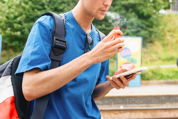 Young man using vape and smartphone at train station in the afternoon