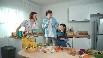 Caucasian skilled father, mother and asian daughter making breakfast while dancing together. Skilled mom playing ukulele while preparing vegetable at modern kitchen. Healthy food concept. Pedagogy.