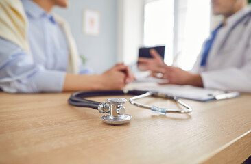 Close up shot of a stethoscope on a wooden table, with a doctor and patient talking in a blurred...