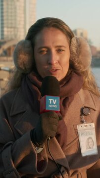 Medium vertical shot of Caucasian female journalist in winter coat, earmuff headphones speaking into microphone while reporting from city street for local TV channel