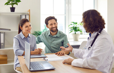 Smiling happy young couple husband and his wife sitting at the desk and talking with a male family doctor therapist in medical clinic or hospital planning pregnancy. Health care and check up concept.