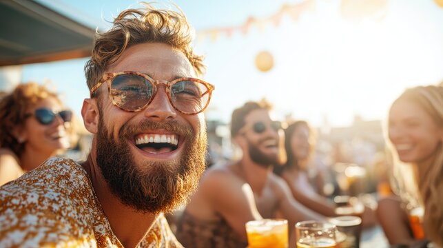 A joyful man with a beard smiles broadly while sitting with friends around a table filled with drinks during sunset, capturing moments of laughter and connection.
