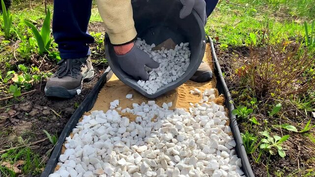 Man worker strewing white decorative split gravel, stones,rocks on path,road,walkway,pathway made of sand on flower bed lawn in garden, landscape design.