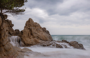 A rocky shoreline with a waterfall, creating a serene and peaceful atmosphere. The waterfall is...