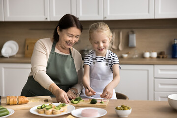 Loving granny teach to cook little grandkid girl, wear aprons stand at dining table cutting fresh cucumbers, preparing delicious sandwiches for breakfast. Older gen teaching to younger, share skills