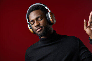 A young Black man enjoying music with headphones, wearing a stylish black turtleneck on a rich red background