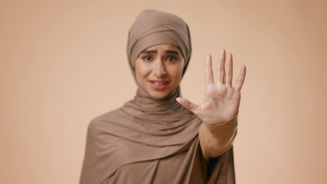 A young Middle Eastern woman wearing a hijab stands against a plain background, raising her hand in a stop gesture to convey a message. She appears focused and serious in her expression.