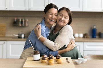 Fototapeta premium Young adult daughter cuddling tightly her older smiling mother, women wear aprons standing together, hugging, while cooking together sweet chocolate muffins, family recipe dessert on weekend at home