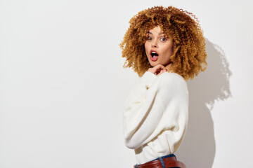 surprised woman with curly hair, white sweater, standing against a plain background, playful...
