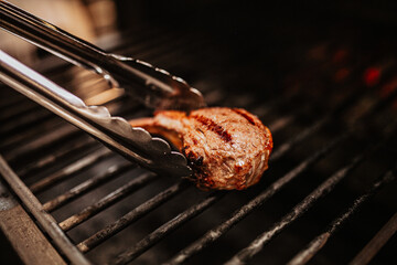 A grilled meat chop resting on hot metal grates, with char marks and steam rising. Tongs hover nearby in a rustic kitchen environment. High quality photo