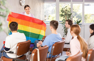 Obraz premium Smiling young female student talking about international LGBT social movements during lecture in auditorium, showing colorful rainbow flag