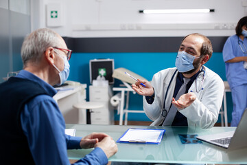 Doctor in lab coat talks to senior patient, both wearing face masks, reviewing symptoms and medical treatment during clinic checkup. Physician with clipboard nearby is giving health advice to old man.