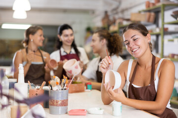 During master class, female participant draws pattern on walls of white ceramic bowl with brush. Girl paints blank, makes individual order. Hand made art, creative pastime.