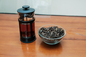 A French press with hot tea inside and dry tea on glass bowl on the wooden table. Concept of...
