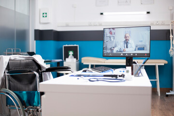 Vacant clinic room with desktop pc showing male physician conducting online consultation. Empty medical office for doctor in wheelchair shows growing role of technology in remote healthcare services.