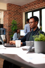 Black businessman seated at desk, using his digital device to reply to work emails and review daily tasks. Male entrepreneur typing on his laptop, preparing startup business project in modern office.