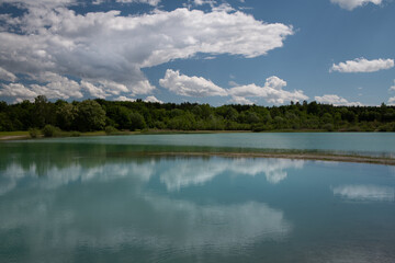 Fototapeta premium The blue sky is reflected in a lake in Bavaria and the clouds are reflected in it. In the background is a group of trees
