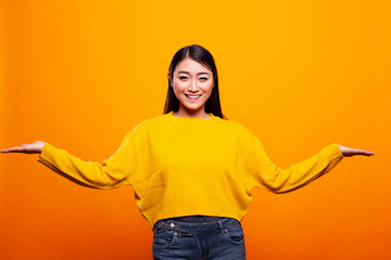Beautiful woman smiles happily, using hand gestures to compare two options, against yellow background. Asian lady cheerfully demonstrating different objects on open palms, presenting and advertising.