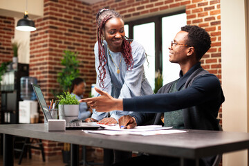 Smiling black woman brainstorming marketing plans with colleague seated at desk with laptop in startup office. African american employees collaborating and sharing strategy plans in brick wall room.