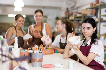 Smiling young girl in apron painting on handmade ceramic bowl with brush in pottery workshop