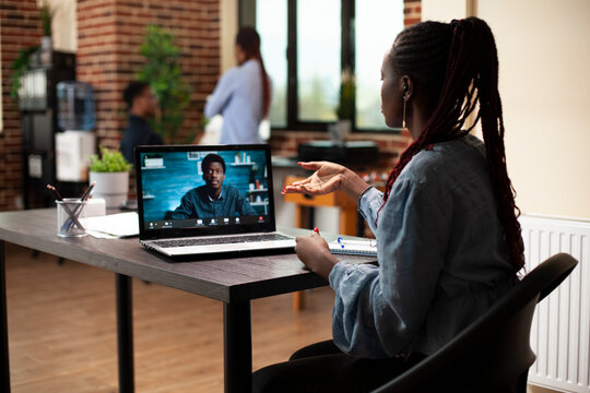 Black woman uses laptop to collaborate remotely, discussing project plans with coworker in virtual meeting. Female advisor at desk engaging in video call with male client to share marketing strategies
