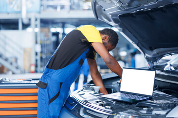 Mockup laptop on car with hood open while african american repairman in blurry background replaces motor. Isolated screen device next to adept garage professional fixing customer automobile