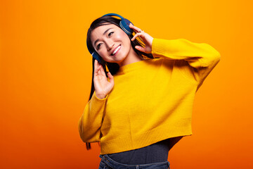 Asian lady with a joyful expression, enjoying music on wireless headphones against a vibrant backdrop. Smiling young woman in a yellow pullover listens to her favorite audio on a digital gadget.