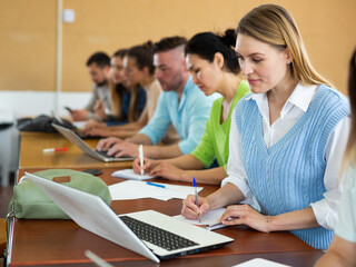 Portrait of a young girl student studying on a laptop and writing synopsis in a copybook during classes in a university ..auditorium with fellow students