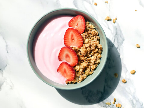 A vibrant bowl of strawberry yogurt mixed with crispy granola, presenting a visually appealing and healthy snack choice. Top view, white marble table.